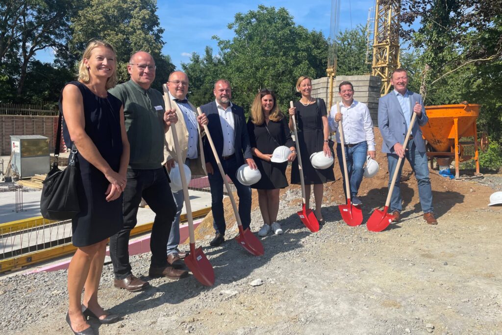 Gruppenfoto bei Spatenstich auf Baustelle mit roter Schaufel.