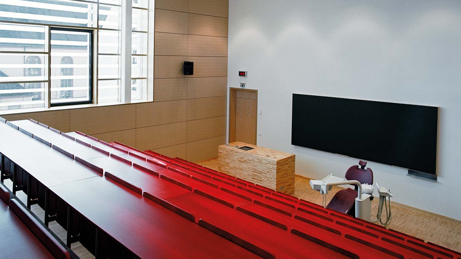 Leerer Hörsaal mit roten Sitzreihen und großem Bildschirm.