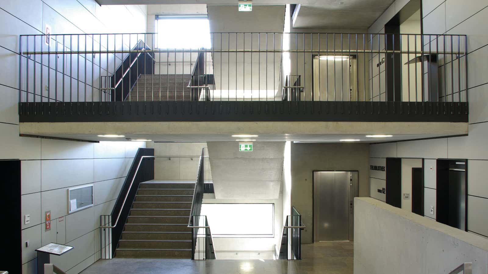 Modernes Treppenhaus mit Aufzug und Notausgangsschildern.